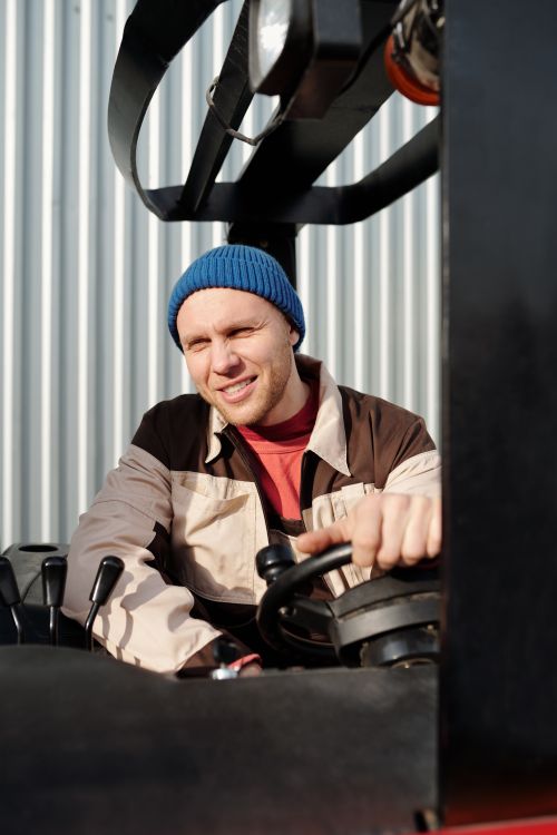Free Smiling man wearing beanie operating a forklift in a sunny outdoor setting. Stock Photo