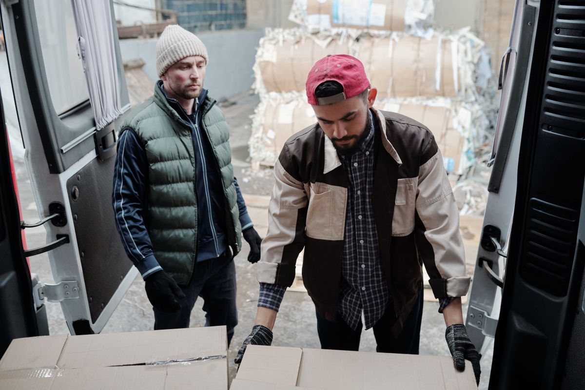 Two men loading cardboard boxes into a delivery van at an urban site.