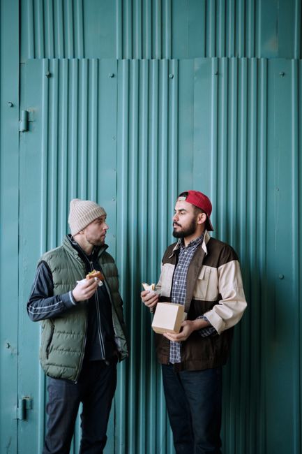 Two men enjoy a casual conversation and meal in front of a textured green wall, captured in an outdoor setting.