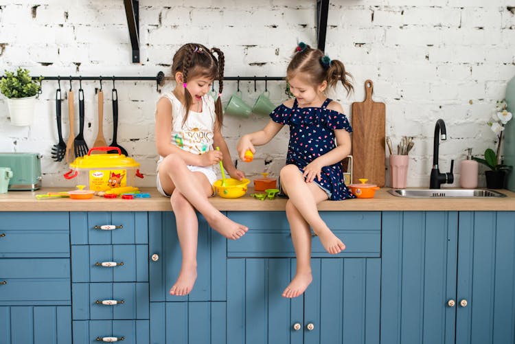 Two Girls Sitting On A Kitchen Counter Playing With Plastic Toys