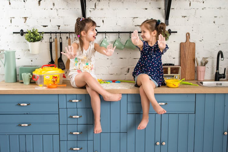 Two Girls Sitting On A Kitchen Counter With Arms Raised