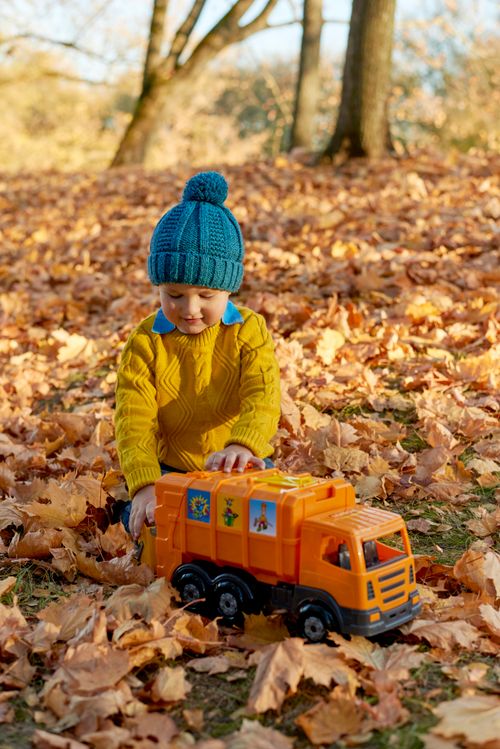 Free A young boy in a blue hat and yellow sweater plays with an orange toy truck amidst fall leaves. Stock Photo