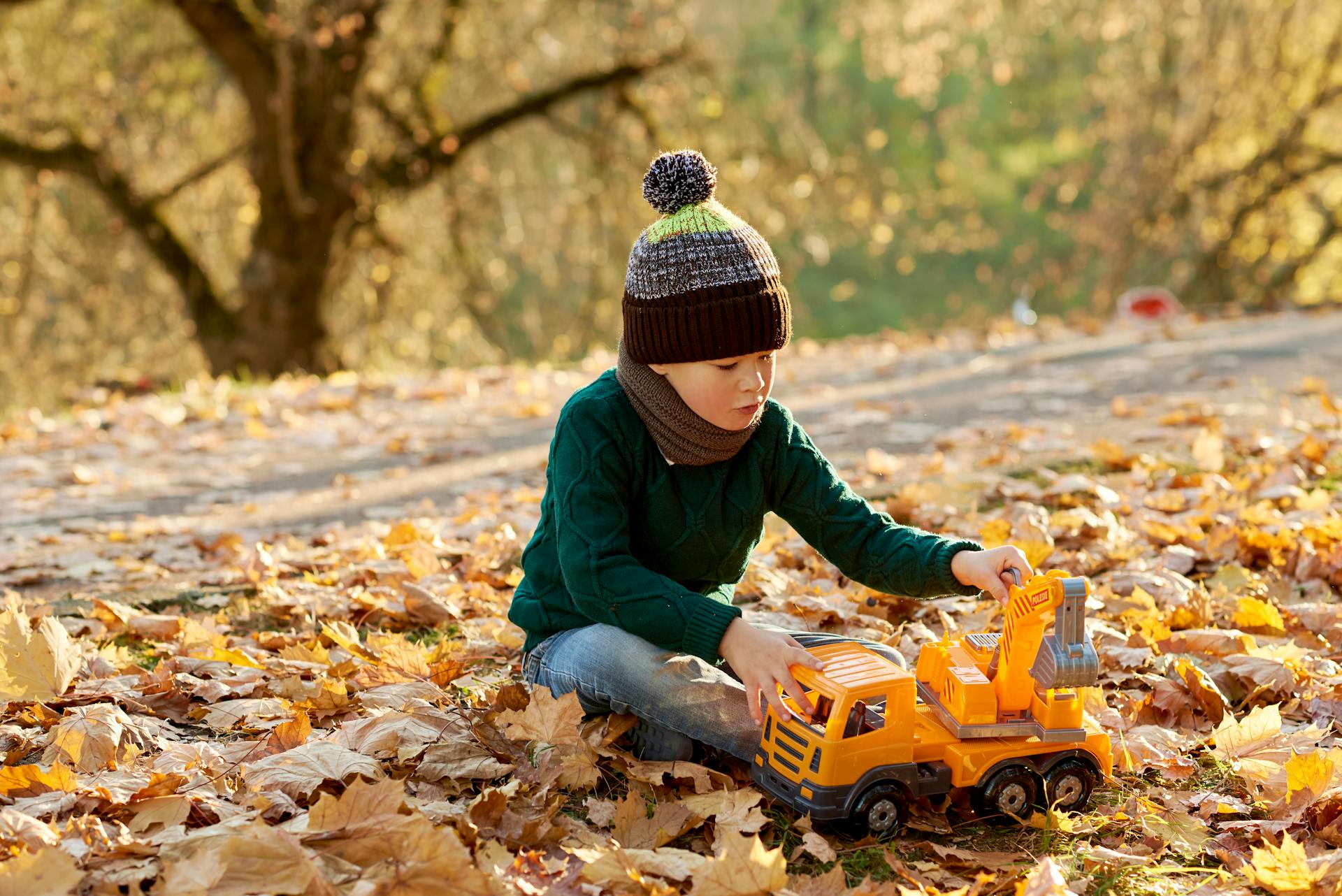 A young boy in fall clothing plays with a toy excavator amidst colorful autumn leaves.