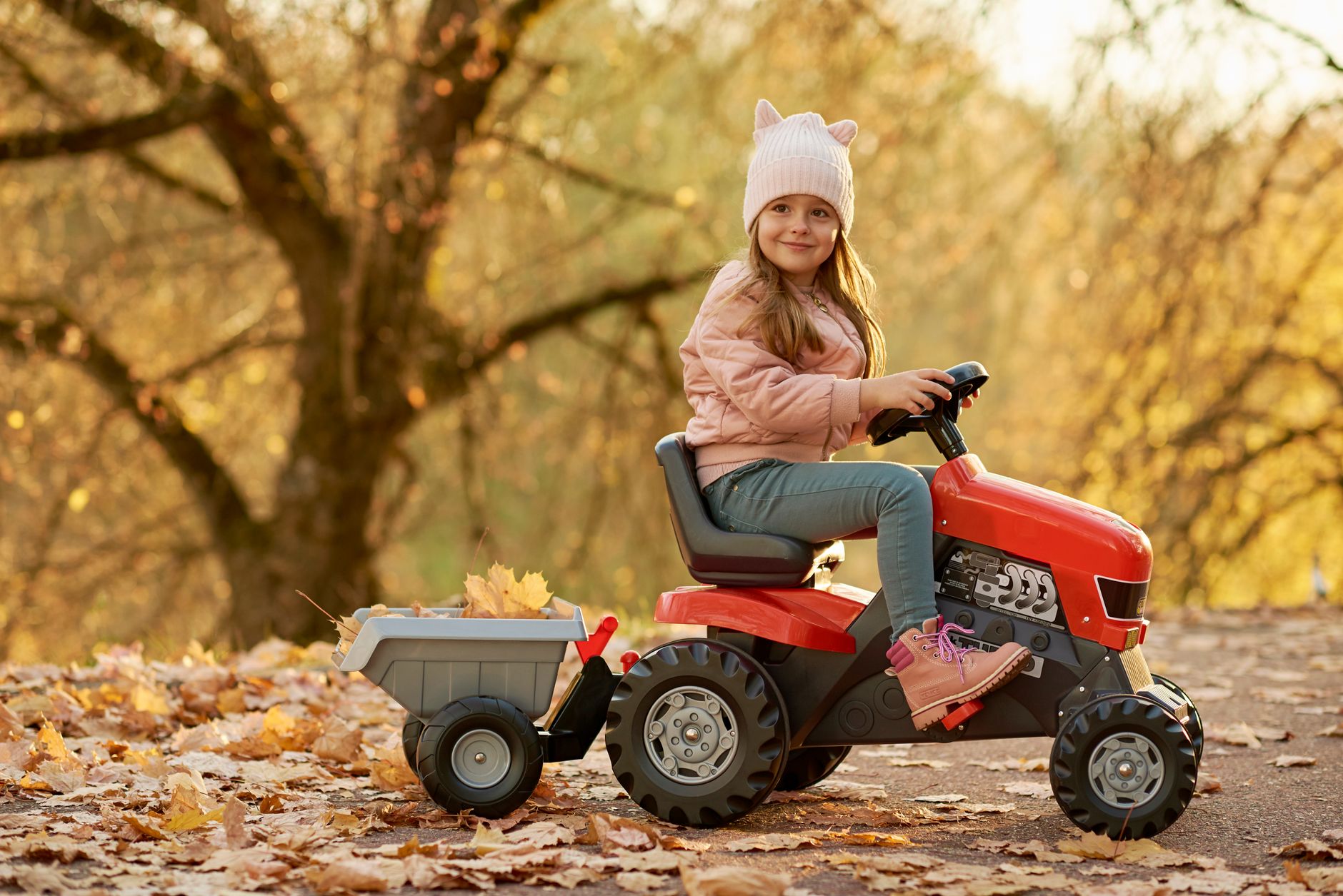 A girl smiles while riding a toy tractor in a park filled with autumn leaves.