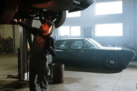 Mechanic working under a lifted car in an indoor garage setting with vintage vehicle in background.