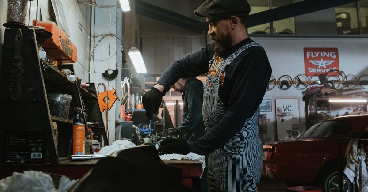 Mechanic performing repairs in a vintage garage setting, using tools and machinery.