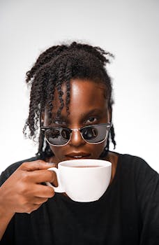 Stylish woman with curly hair and sunglasses holding a white mug against a white background.