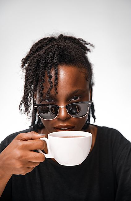 Stylish woman with curly hair and sunglasses holding a white mug against a white background.
