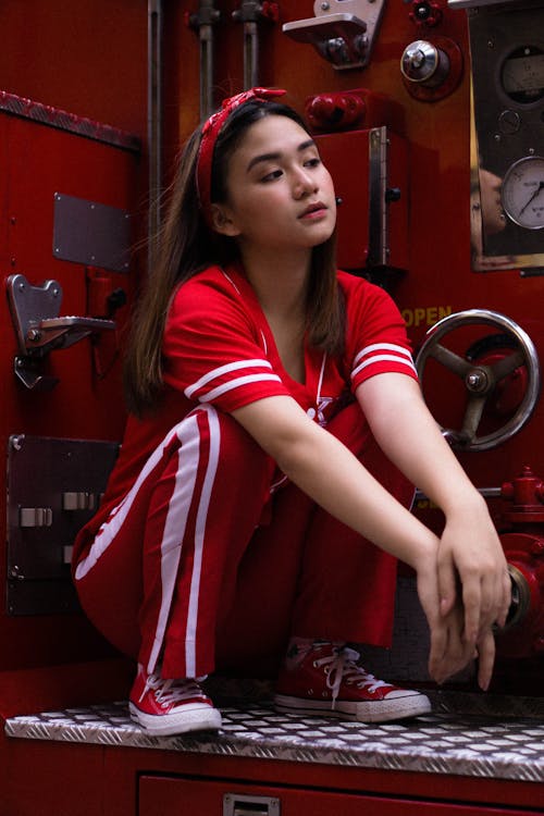 Full body young Asian female wearing red activewear and hairband hunkering down on counter in red machinery room while looking away arrogantly