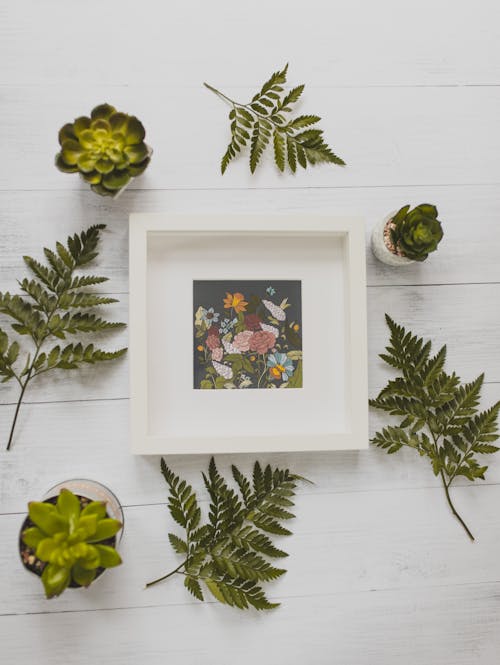 Free Top view composition of white square frame with floral picture placed on white wooden desk amidst green lush succulents and plant leaves Stock Photo