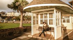Full body young female in casual warm wear sitting on bench in white small summerhouse located in green district of southern town