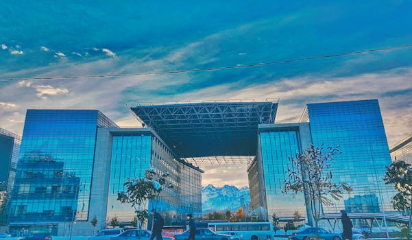 Contemporary glass skyscraper with mountains in Almaty framed by blue skies.