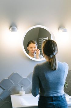 A woman uses a mirror to apply cream as part of her skincare routine.