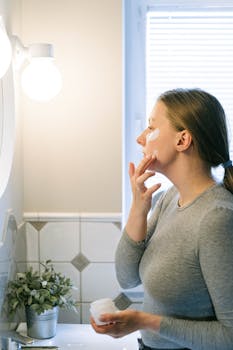 A woman applying face cream in a well-lit bathroom, focusing on self-care and skincare routine.