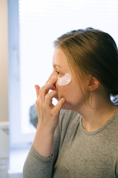Free Caucasian woman in gray shirt applies under-eye patch for skincare in natural light. Stock Photo