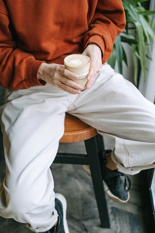 Free A Person in Brown Sweater and White Pants Holding a Drinking Glass while Sitting on a Wooden Stool Stock Photo