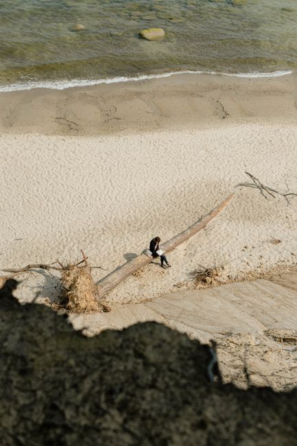 A lone woman sits on a log by the beach, embracing solitude under the bright summer sky.