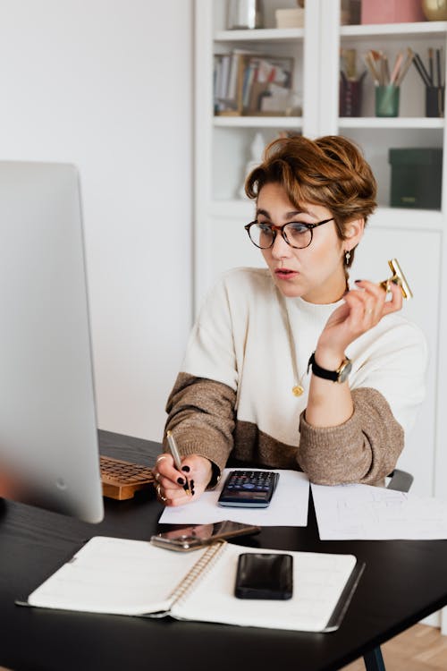 Free Surprising adult short haired female employee in eyeglasses reading unexpected information on computer and taking notes Stock Photo