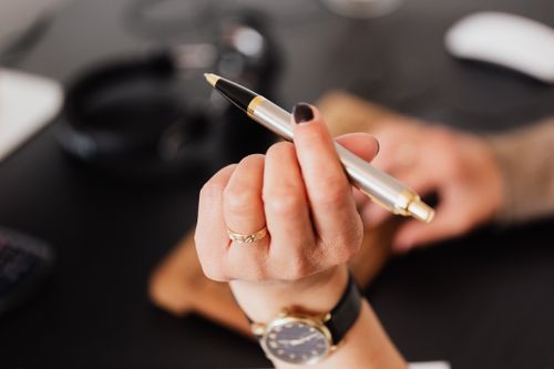 Free A close-up of a hand holding a silver pen, set against a blurred office background. Stock Photo