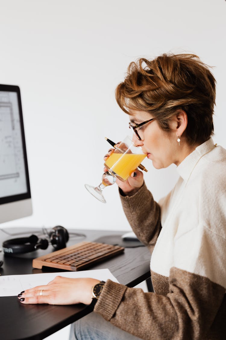 Focused Female Drinking Refreshing Juice While Working At Table In Office