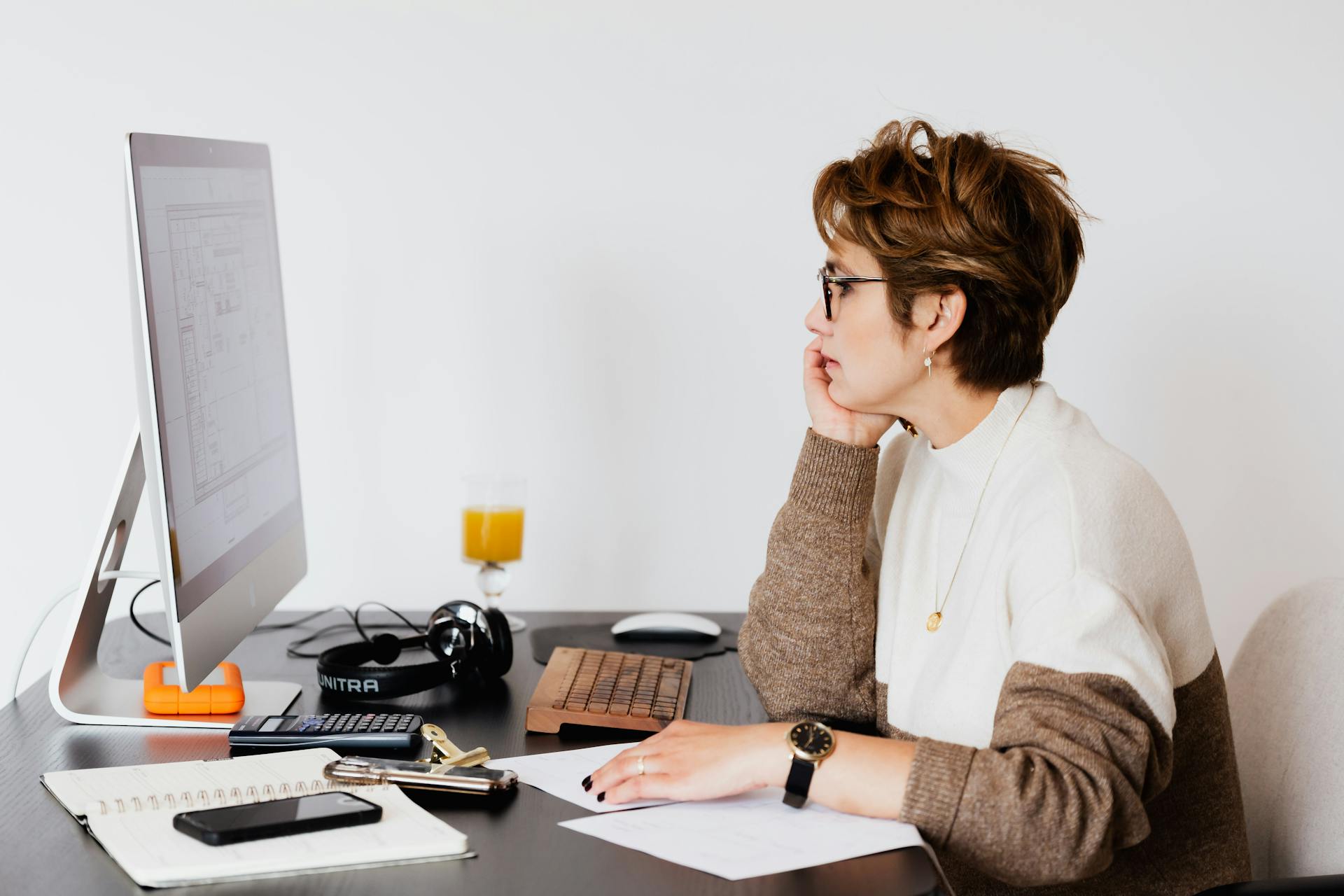 Side view of elegant female architect in glasses sitting at desk with modern PC and studying plan of building on computer screen