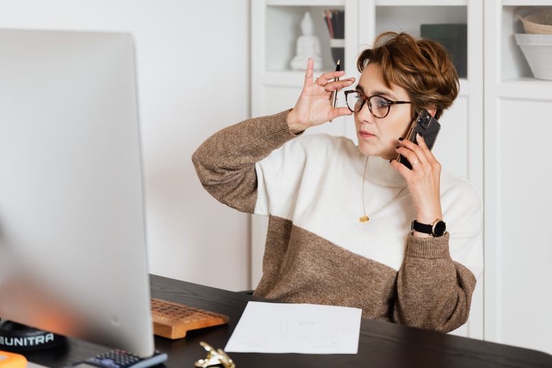 A woman in glasses taking a phone call at her desk, one hand raised mid-gesture, with her monitor turned away.