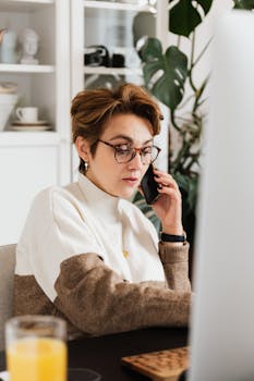 Strict female director talking on mobile phone while working on computer at desk with orange juice in homelike office