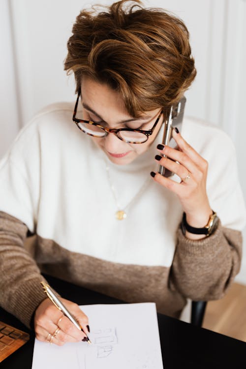 From above of serious female interior decorator in eyeglasses talking on mobile with client and writing down details of new project