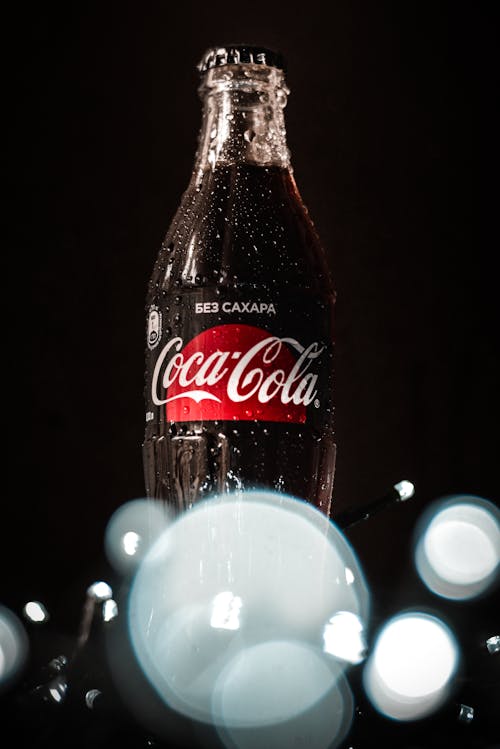 Low angle wet glass bottle of fizzy soda drink placed on table in dark room near fairy lights