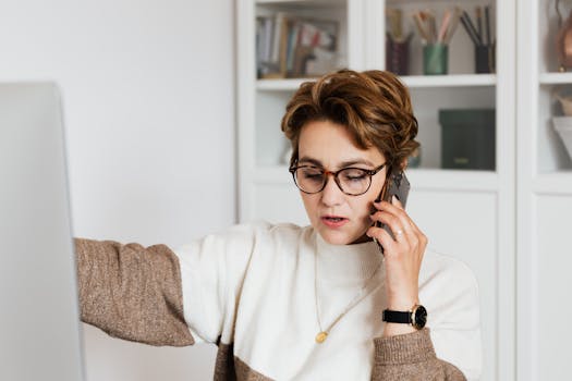 Female remote worker in eyeglasses talking on mobile with boss while working on project at home