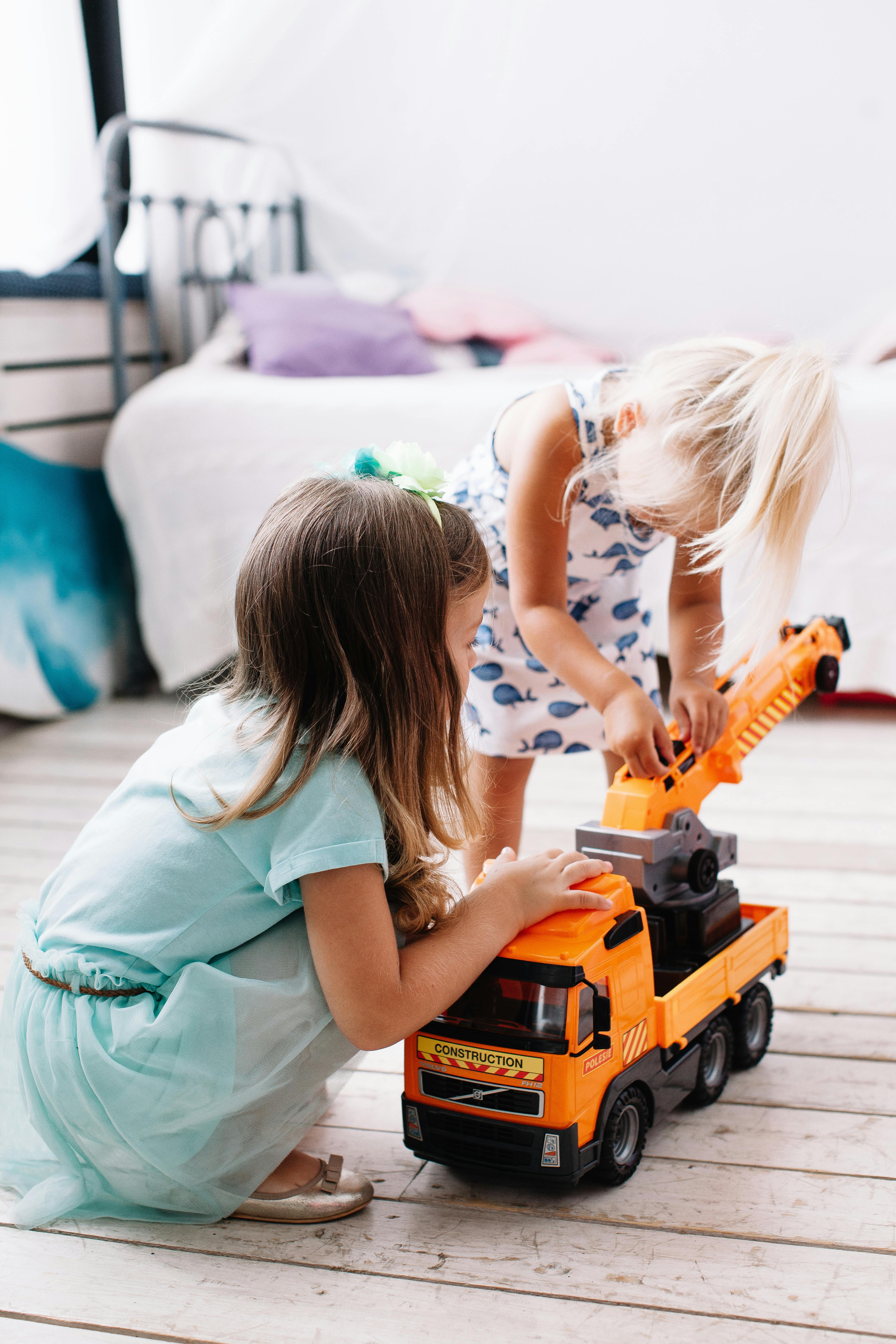 Two young girls playing with a toy truck on the floor of a bright and cozy bedroom.