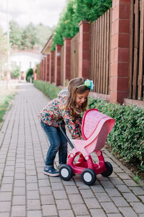 Free Cute girl with floral jacket playing with a pink toy stroller on a stone pavement. Stock Photo