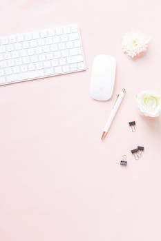 Flat lay of a minimalist workspace with keyboard, mouse, and flowers on a pink background.