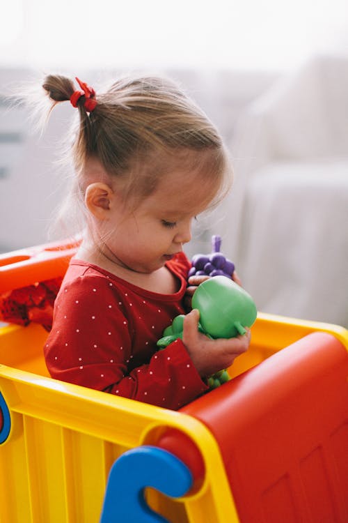 Girl in Red Long Sleeve Shirt Holding Plastic Toys while Sitting on a Toy Truck