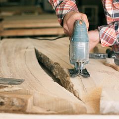 Hand guiding a blue-grey jigsaw along the edge of a live-edge wooden slab, sawdust on the wood