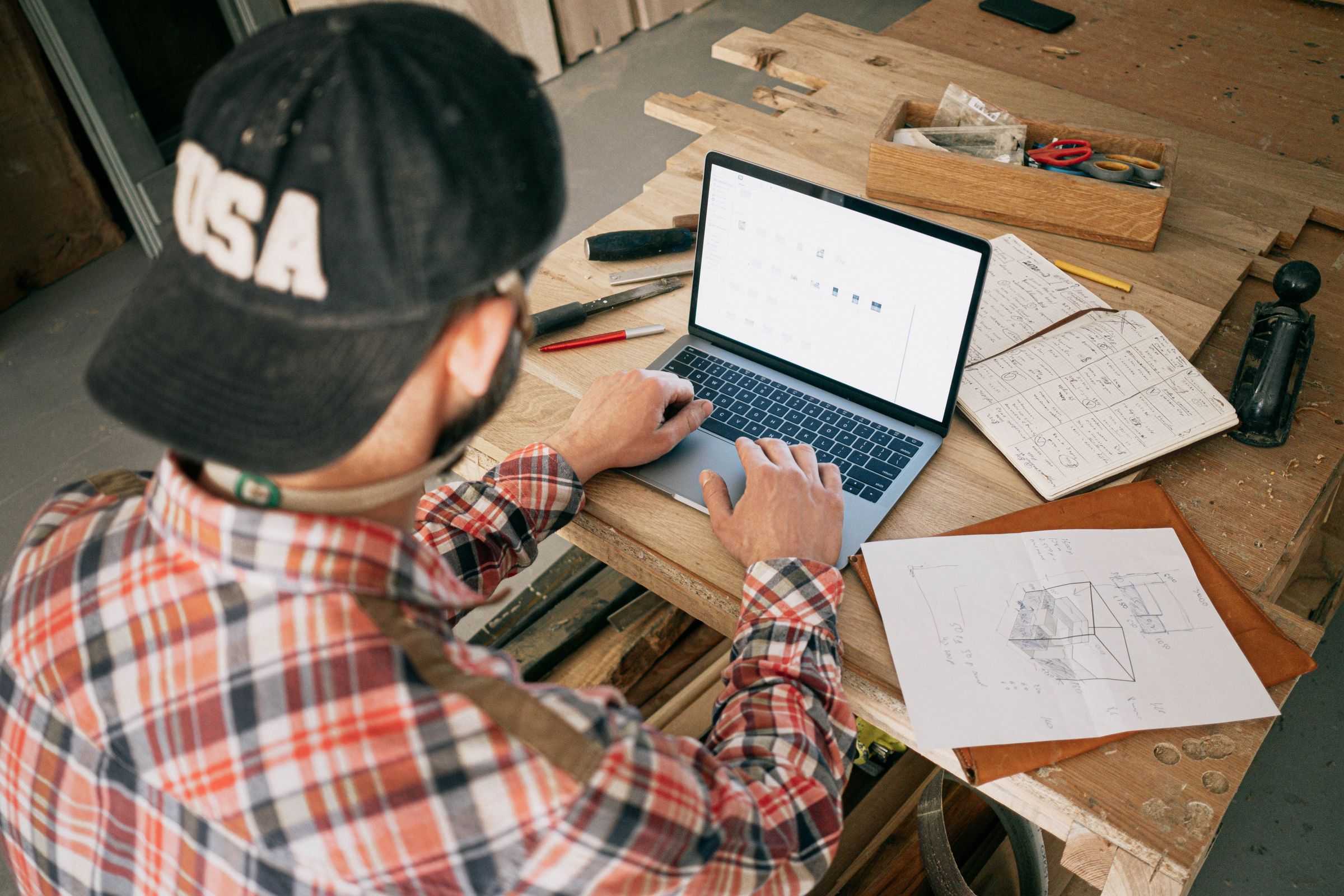 Woodworking business owner using a laptop and project plans at a workshop bench