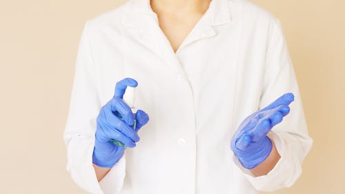 Crop unrecognizable hospital worker in white uniform spraying antiseptic liquid over hands in blue latex gloves