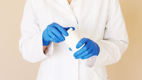 Crop unrecognizable female doctor in white uniform and blue gloves opening plastic white jar of medical pills on light brown background