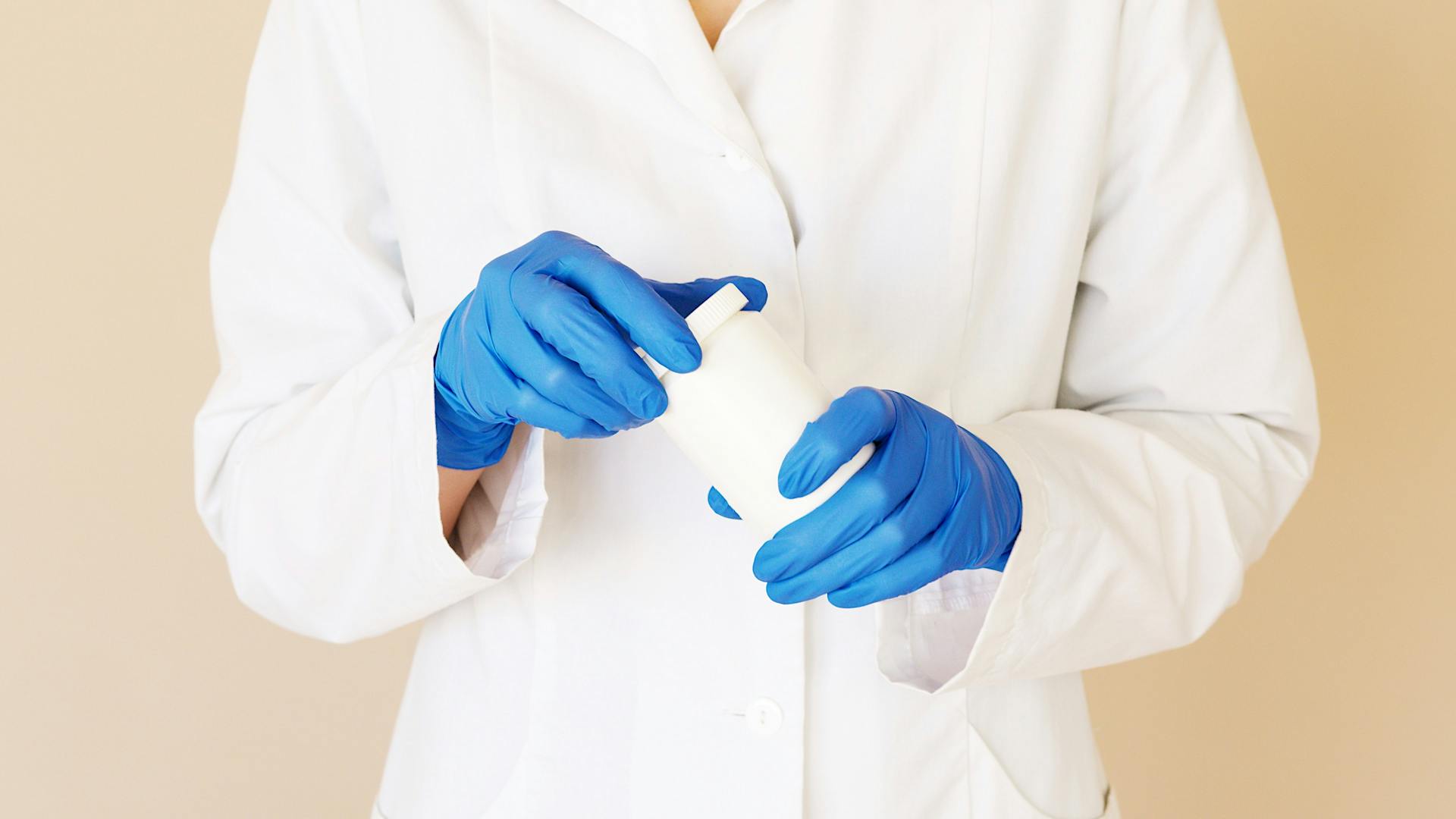 Crop unrecognizable female doctor in white uniform and blue gloves opening plastic white jar of medical pills on light brown background