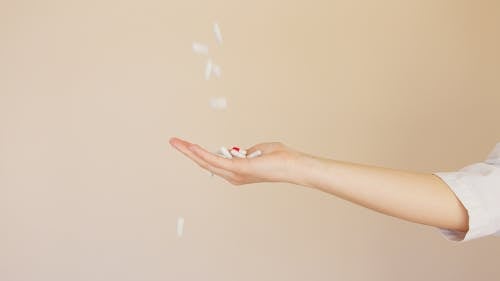 Side view of crop anonymous female pharmaceutist in white coat with palm facing up catching medical capsules falling down from above on light brown background