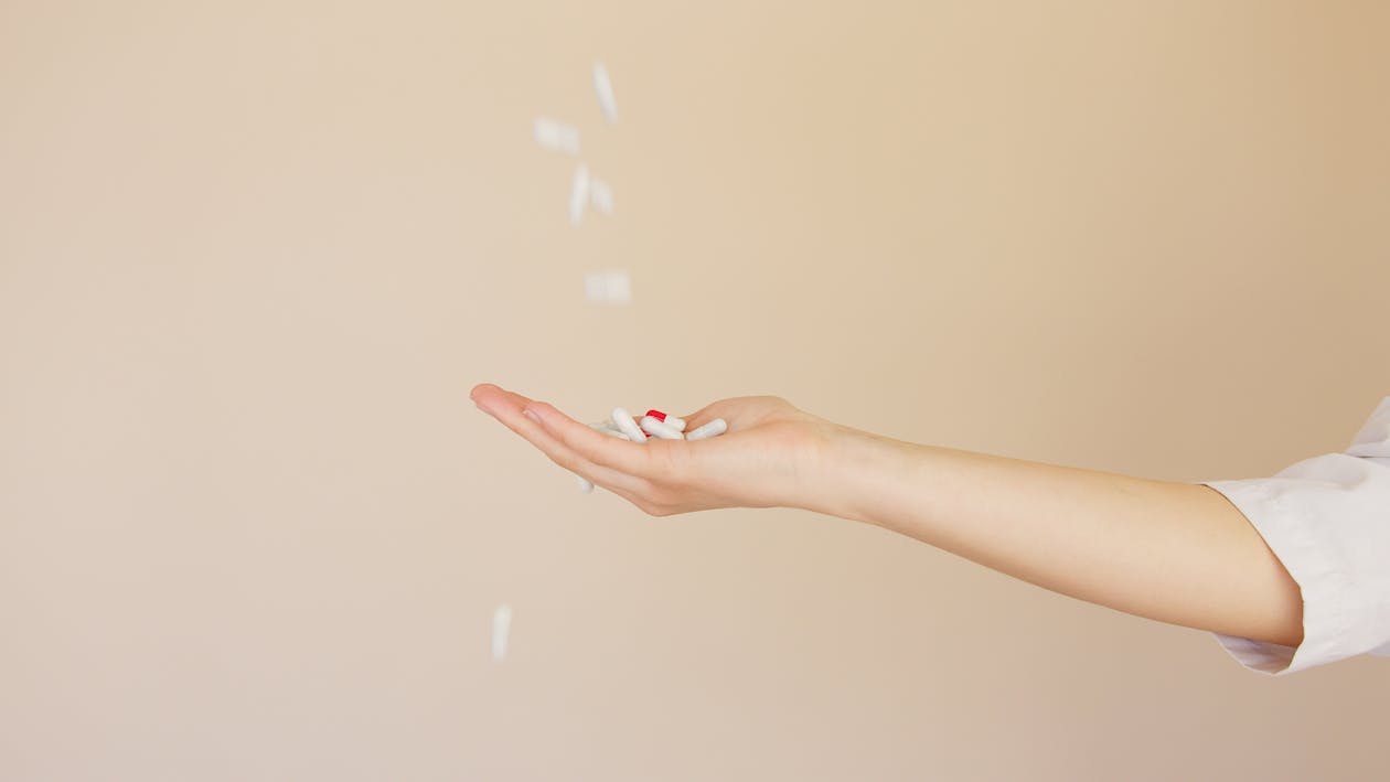 Side view of crop anonymous female pharmaceutist in white coat with palm facing up catching medical capsules falling down from above on light brown background