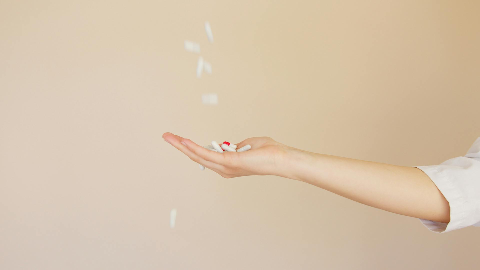 Side view of crop anonymous female pharmaceutist in white coat with palm facing up catching medical capsules falling down from above on light brown background