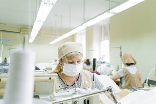Woman focused on sewing in a bright industrial workshop wearing protective gear.