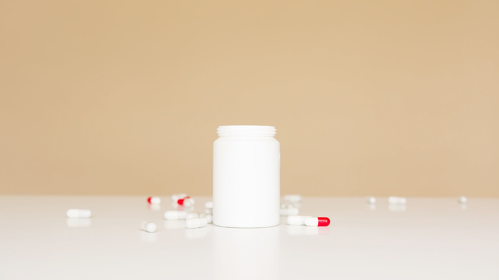 Open white plastic bottle and red and white pills scattered around on white table