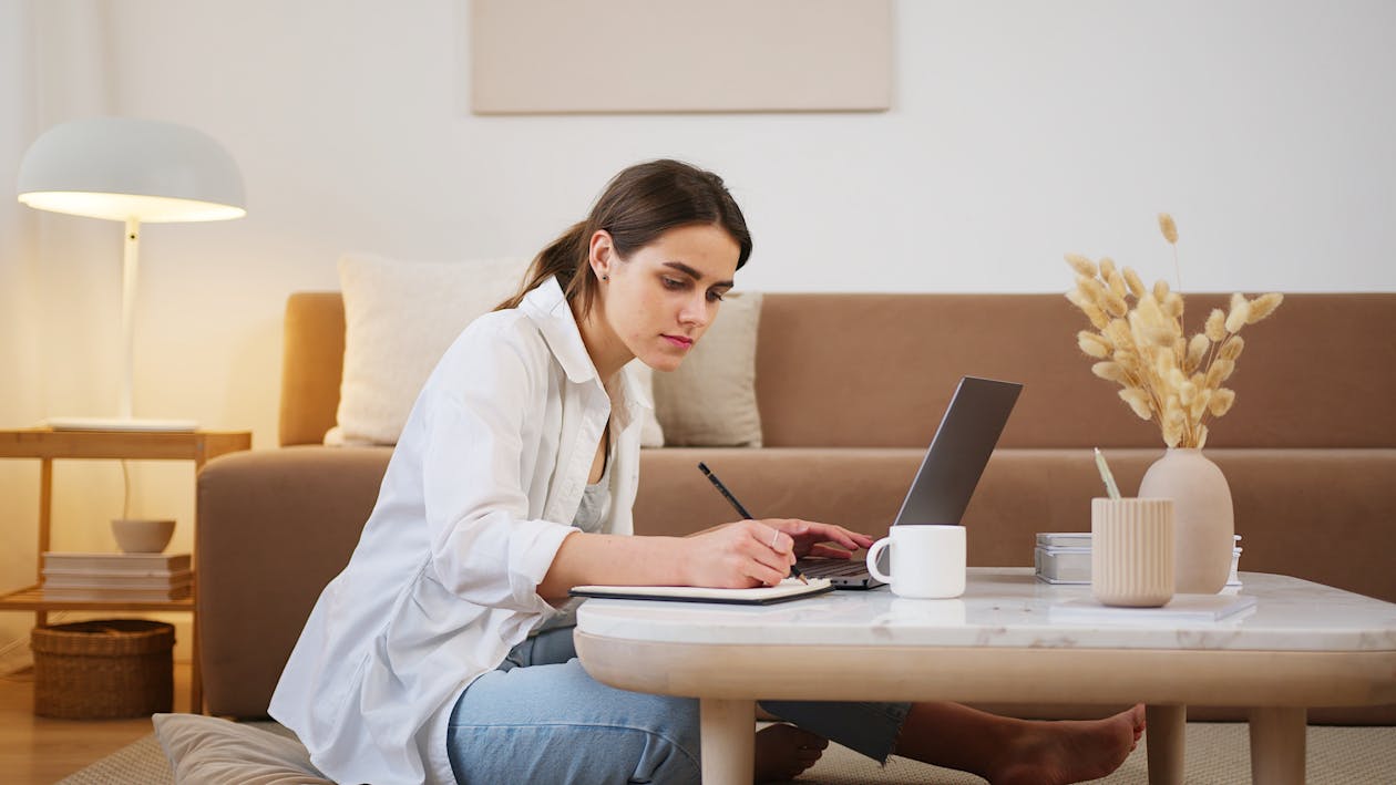 Free Side view full length young focused female in casual clothes browsing modern laptop and writing down notes while sitting on floor in cozy modern apartment Stock Photo