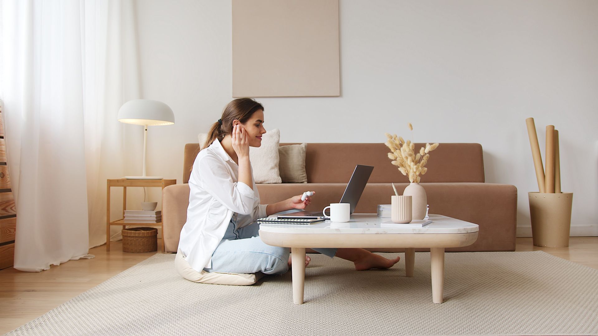 Young woman using laptop with earbuds in a modern living room setting, engaging in a video call.