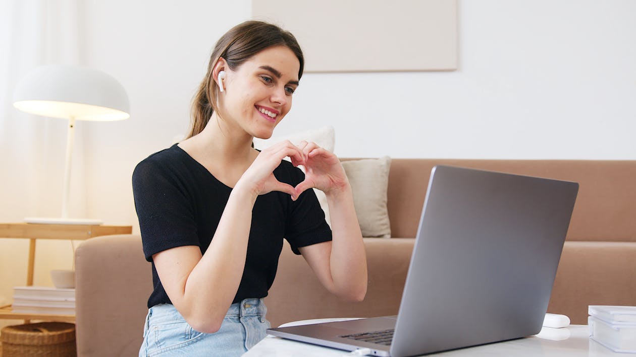 Free Cheerful happy young female in jeans and black shirt having conversation via video call on laptop and making heart shape with fingers while sitting on floor in modern living room Stock Photo