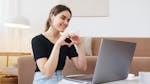 Cheerful happy young female in jeans and black shirt having conversation via video call on laptop and making heart shape with fingers while sitting on floor in modern living room