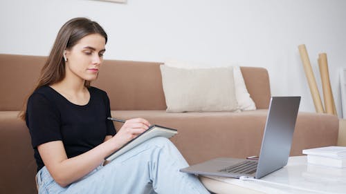 Glad young lady in jeans and black shirt writing down notes while attending online lesson and sitting on floor in cozy modern living room