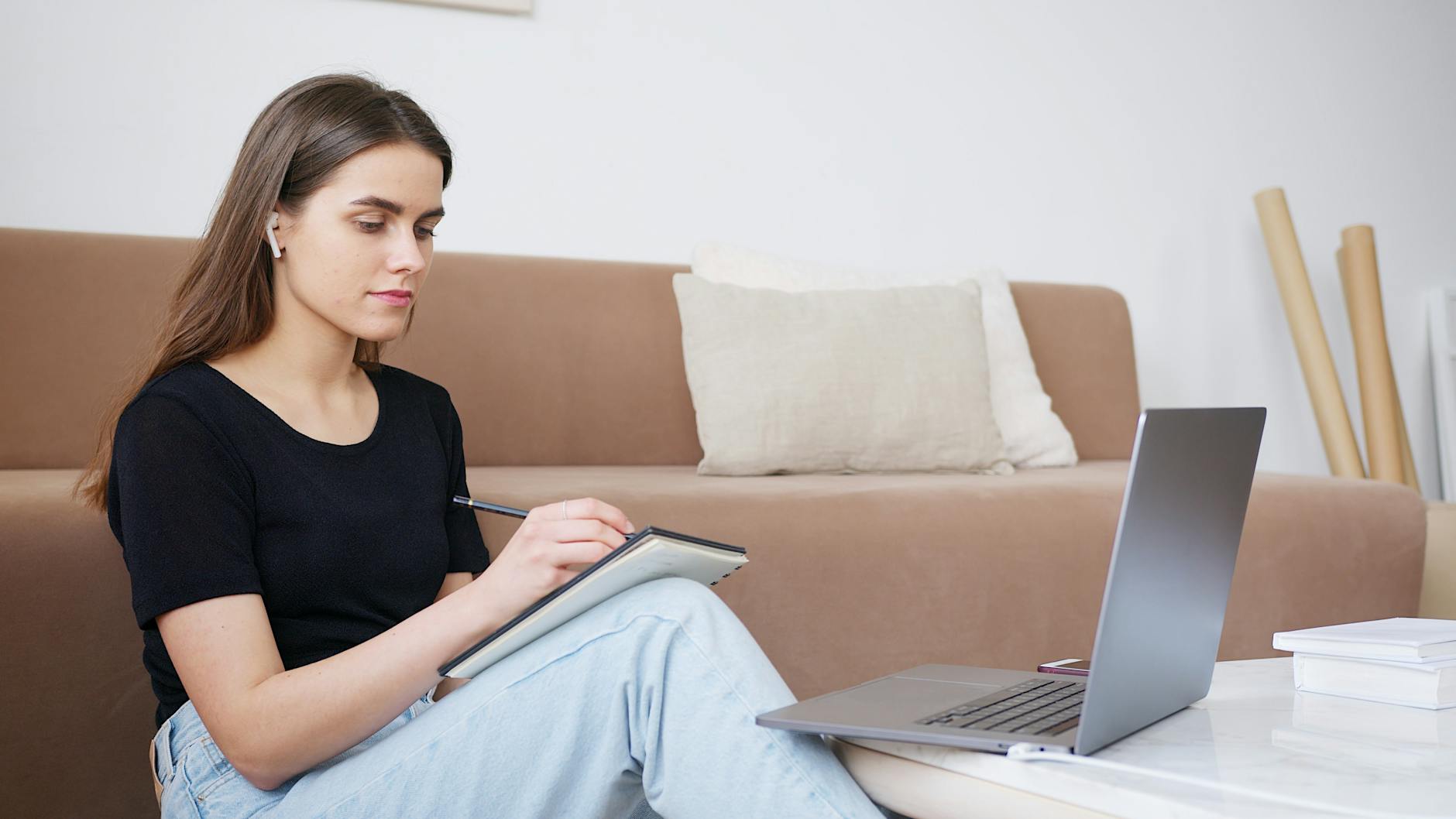 Glad young lady in jeans and black shirt writing down notes while attending online lesson